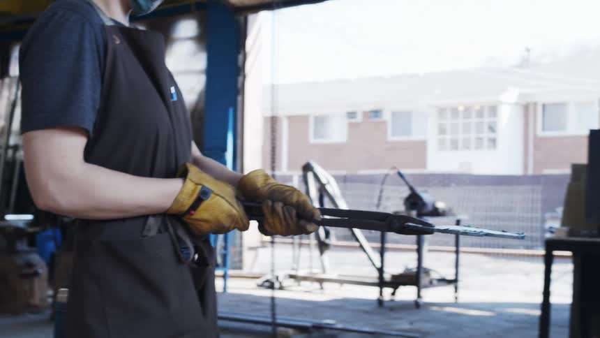 Woman wearing face mask and apron holds metal shape with pincers and carries it across factory for near wide opening showing building next door
