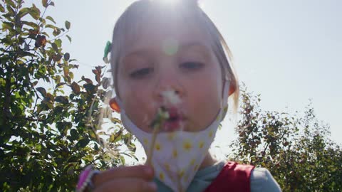 A young girl holds a dandelion up to her mouth and blows its seeds into the wind