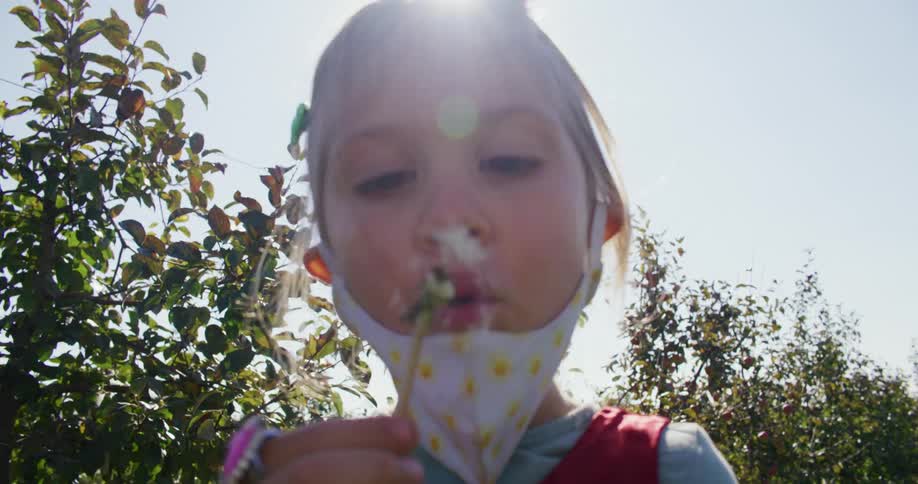 A young girl holds a dandelion up to her mouth and blows its seeds into the wind