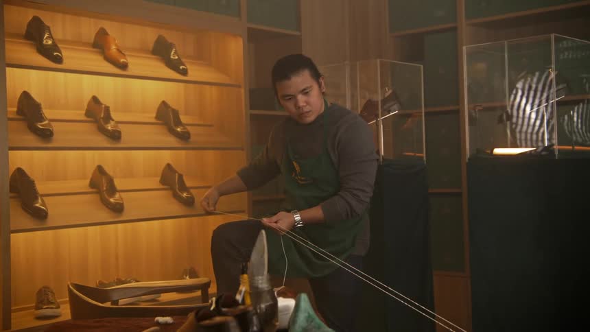 A shoemaker pulls on thread while standing in front of a display of leather shoes