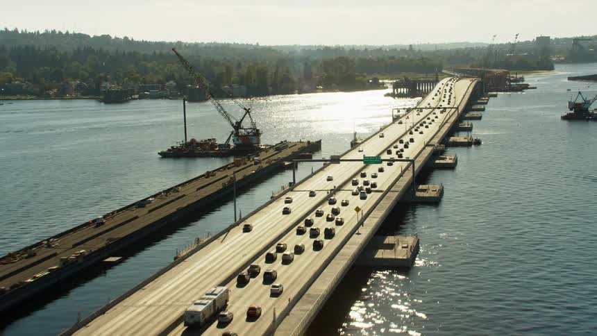 A bridge with vehicles over a river with land the sky and clouds in the background