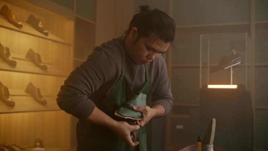 Man stands in workshop wrapping leather to make shoe with display of shoes in background