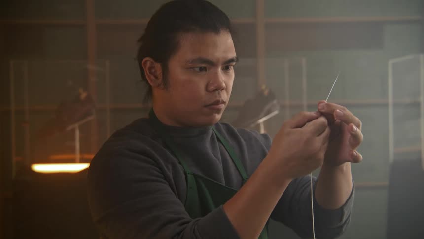 A male shoemaker concentrates as he carefully threads a long needle with string