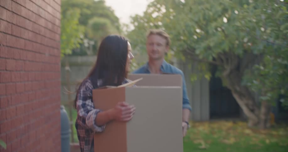 A man and a woman holding boxes and walking by the side of a house with trees the sky and clouds in the background