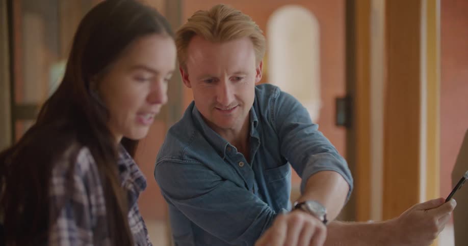 Blonde man with a blue shirt talks to the woman with brown hair and a plaid shirt holding a photo frame in the new house