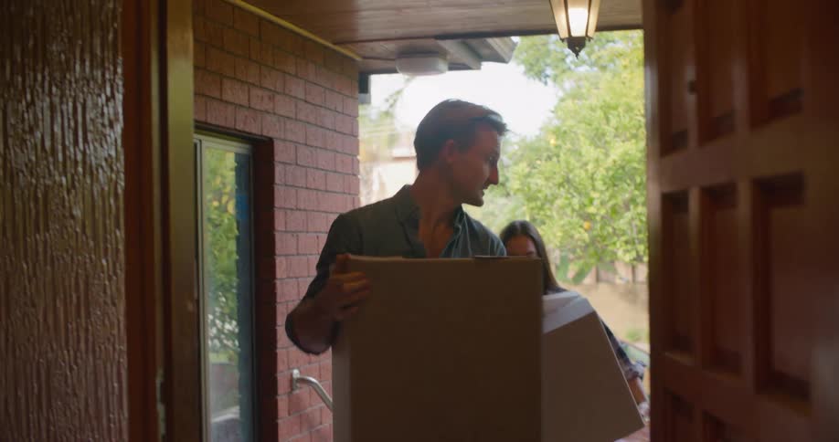 A man and woman walk up steps and through the front door carrying cardboard boxes from the car