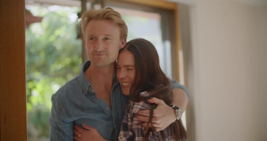 Woman with a plaid shirt and brown hair carries a box into living room and cuddles blonde man with a blue shirt in the new house