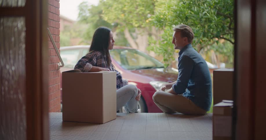A man and a woman sitting on a porch with boxes with a vehicle and trees in the background