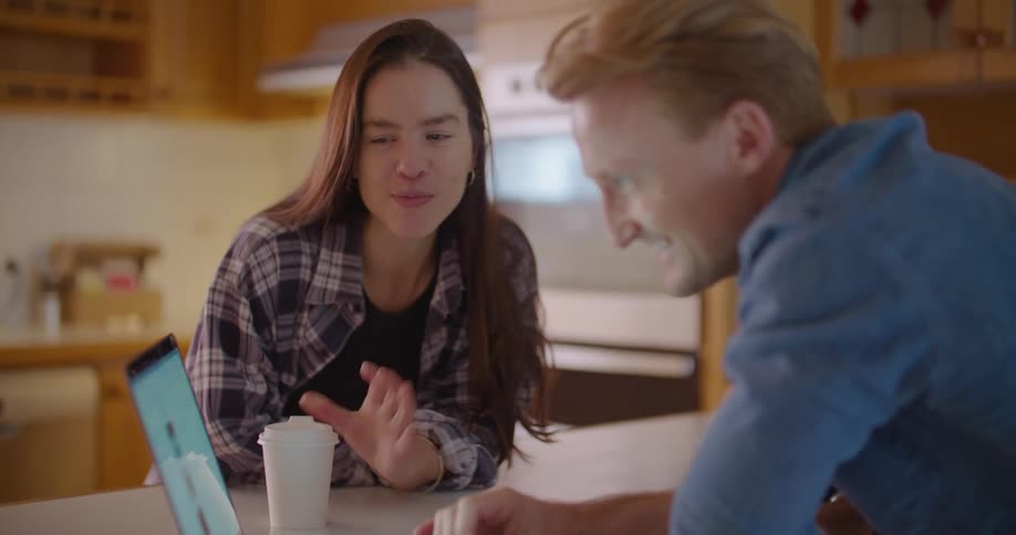 A man and young woman smiling and laughing while using a laptop in a home kitchen