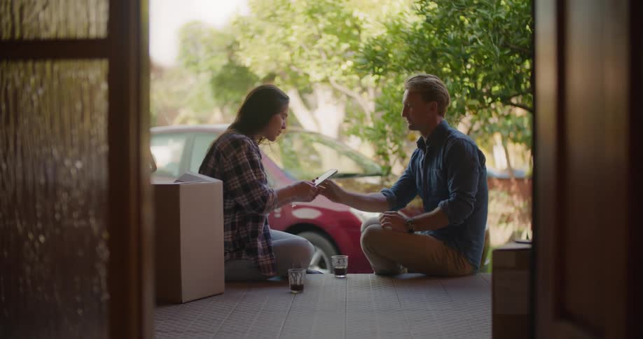 A middle aged man hands a smartphone to a young woman before taking a drink from his glass as they sit together on the porch of a house during the day