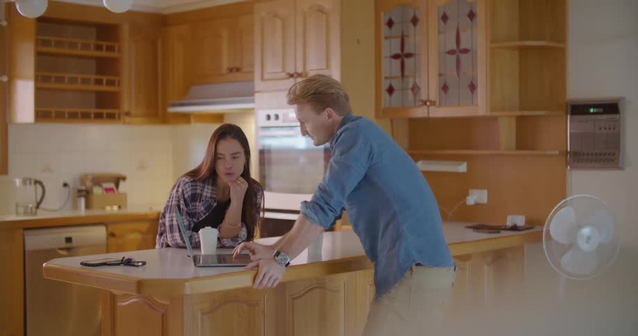 A man leans on a kitchen counter tapping on a tablet as he talks to a client in her kitchen