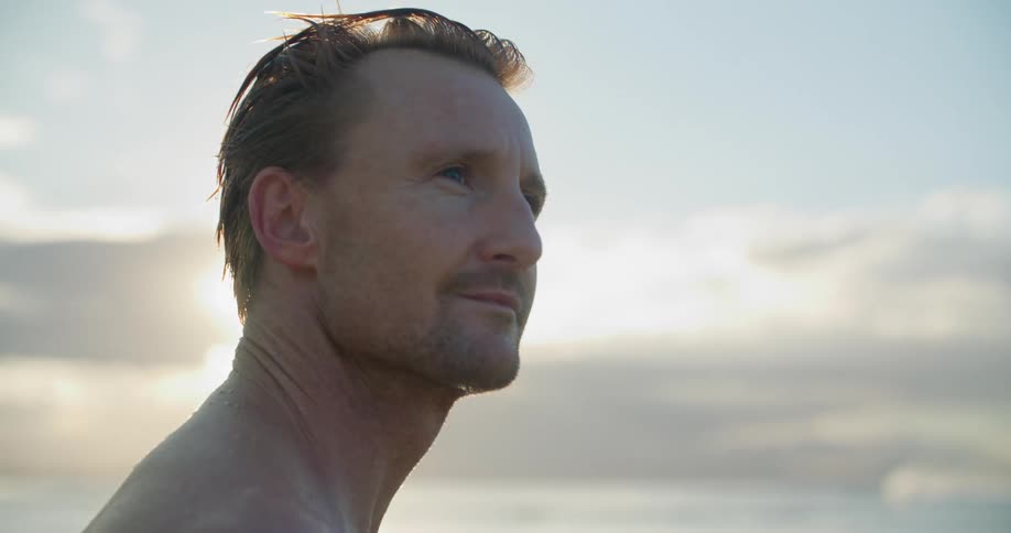 A man smiles softly as he looks around the ocean beach as the sun glares behind him