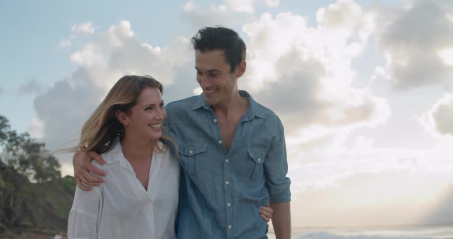 A man and woman smile and talk as they walk the beach with arms around each other