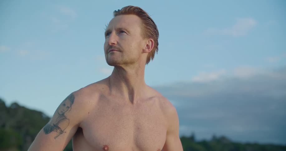 Shirtless blonde man with a tattoo stands and looks around on the beach