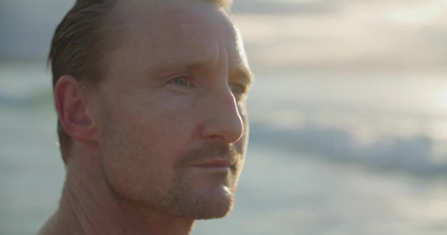 A man standing on a beach smiles as he turns his head to look into the distance on a sunny day