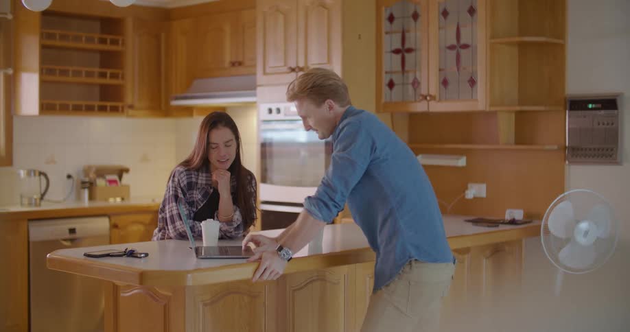 A man scrolls on a tablet and talks with a woman as they stand in a kitchen
