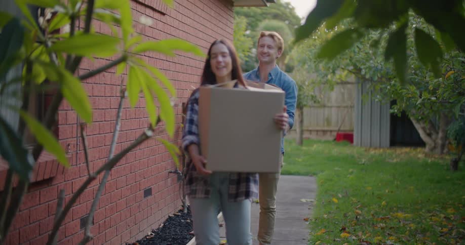 A man and young woman carrying cardboard boxes while walking on a sidewalk next to a brick house