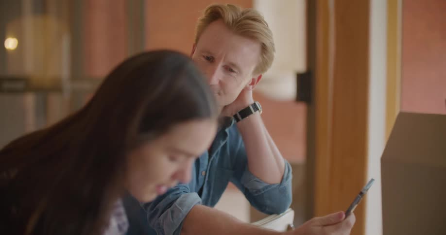 A man shows the screen of a smartphone to a young woman searching a box next to him indoors during the day