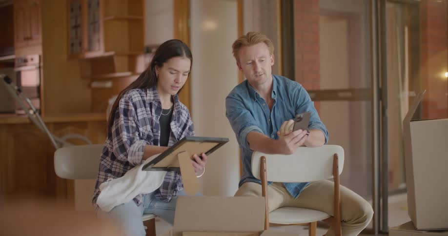 A man scrolls through his phone as his wife unpacks a framed photo from a cardboard box