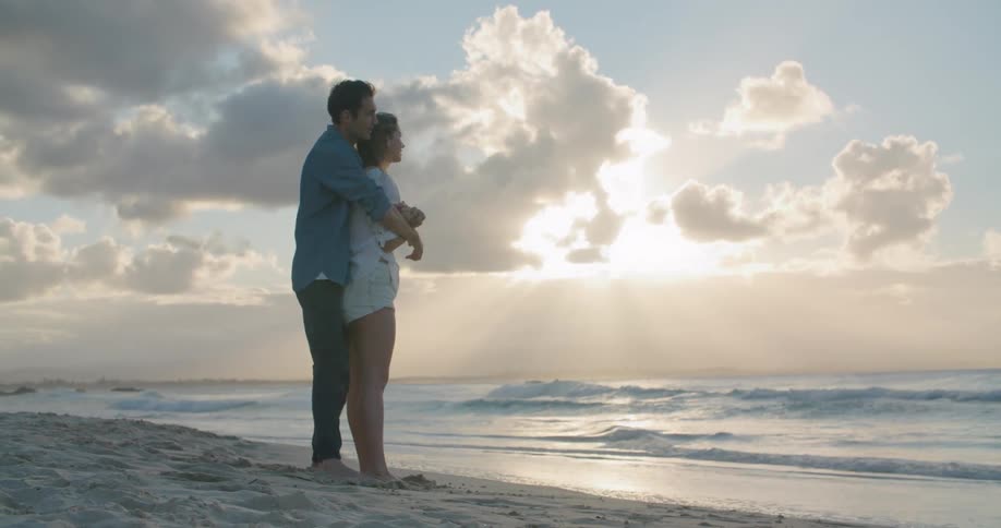 Man in a blue shirt and jeans holds woman wearing a shite shirt on the beach next to the sea