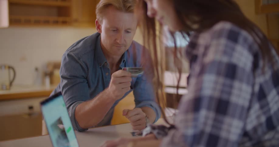 Blonde man with a blue shirt holds up a card and tells woman with a plaid shirt to enter the code to buy furniture on laptop in the kitchen
