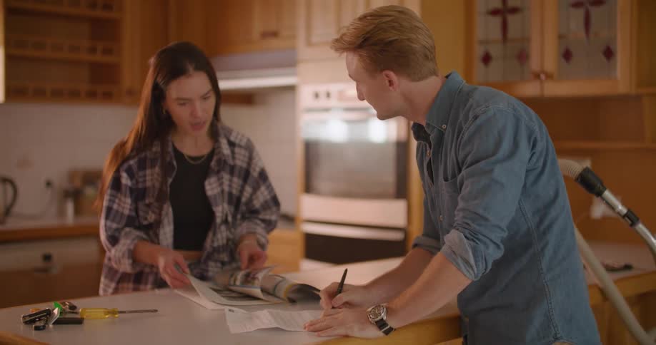 A man and woman stand in a kitchen and talk as she flips through a magazine and he writes on a paper