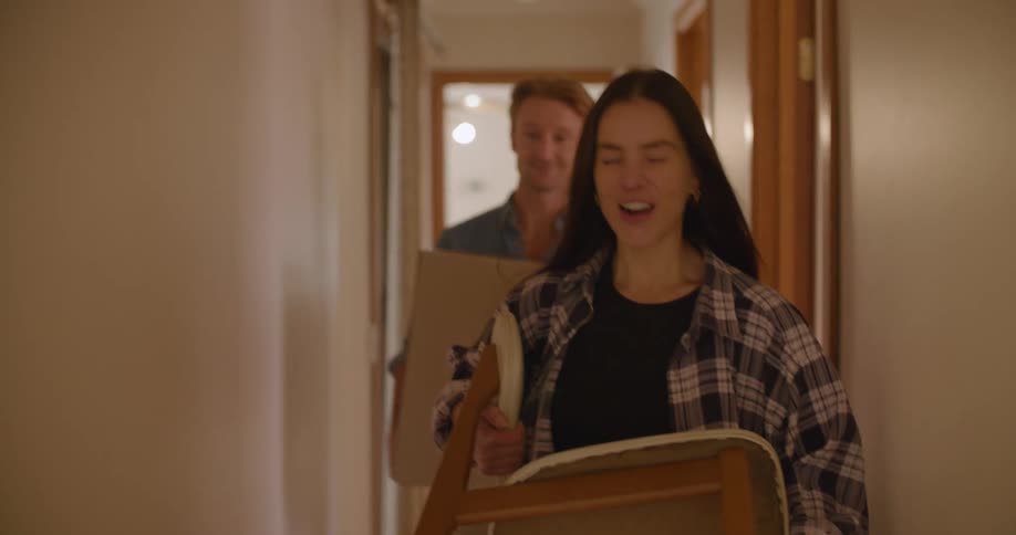 A man carries a box down a hallway and talks and laughs with a woman carrying a chair