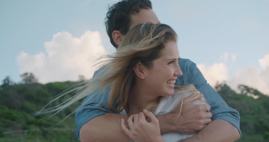 A man hugs his girlfriend from behind and points to the distance as they stand in front of a tall hill