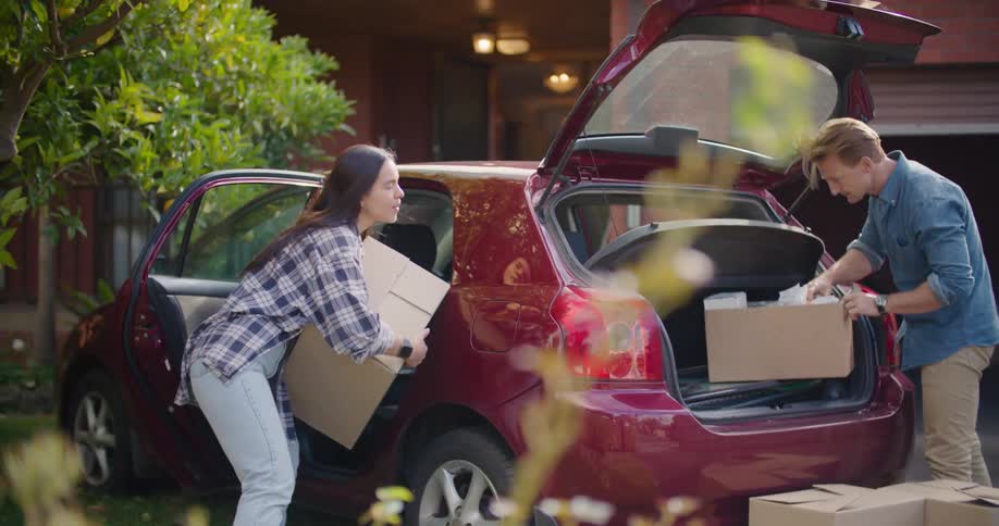 Couple unpacks car of cardboard boxes near building with open garage doors