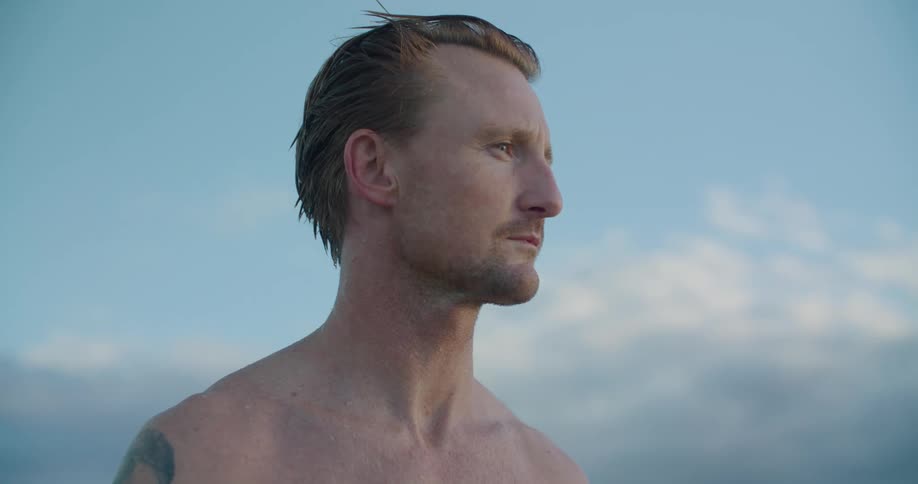 Blonde shirtless man with a tattoo looks around on the beach