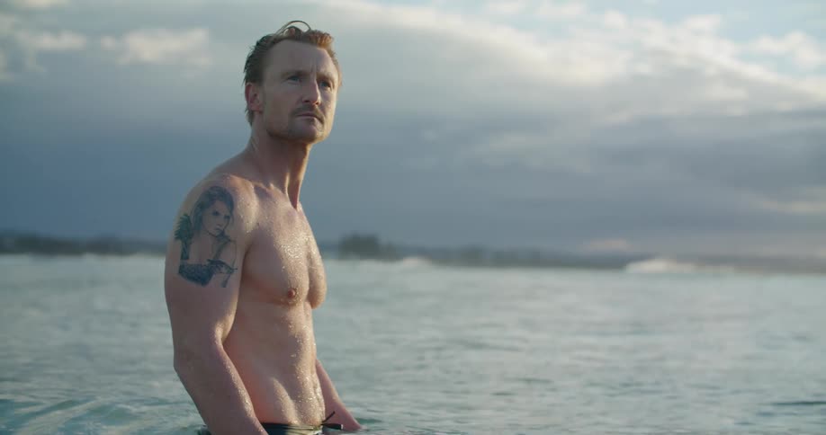 Barechested man stands in water up to his waist looking around as waves roll over surface of ocean under clouded sky