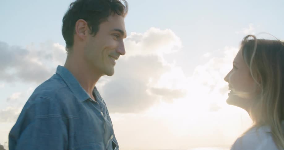 Man with a blue shirt and blonde woman with a white shirt hold hands and kiss each other at sunset on the beach