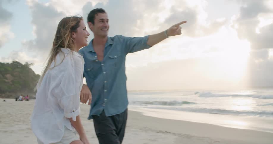 Couple plays around on beach smiling as sun shines through clouds onto waves rolling onto beach