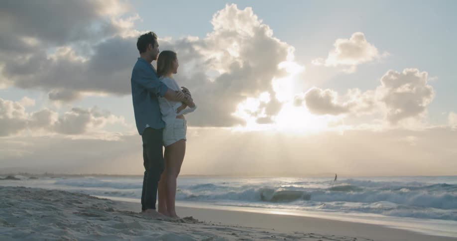 A man hugs his girlfriend from behind as they stand on the beach and watch the ocean