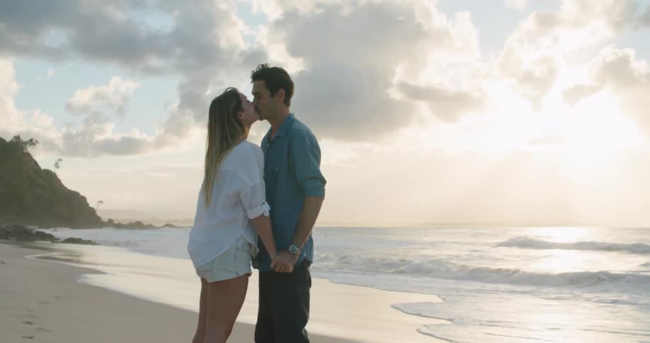 A man and a woman standing on a beach holding hands and kissing with the ocean sky and clouds in the background