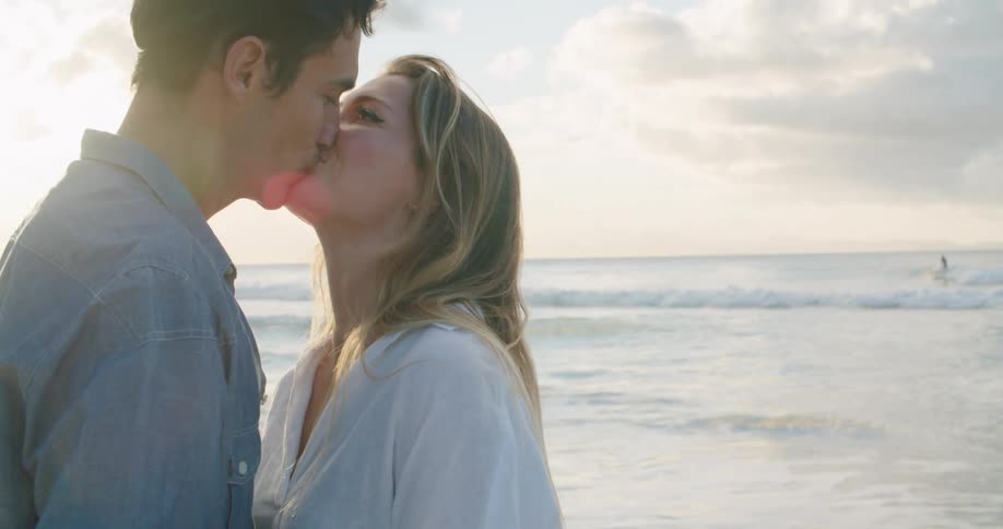 A couple kiss on a beach as a man is surfing in the background