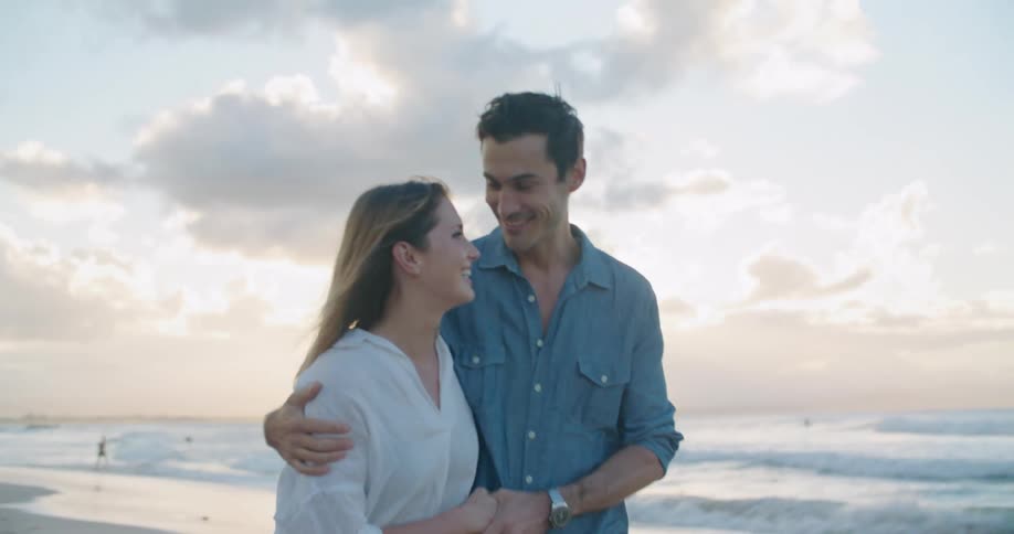 A man and woman walk with arms around each other and holding hands on the beach