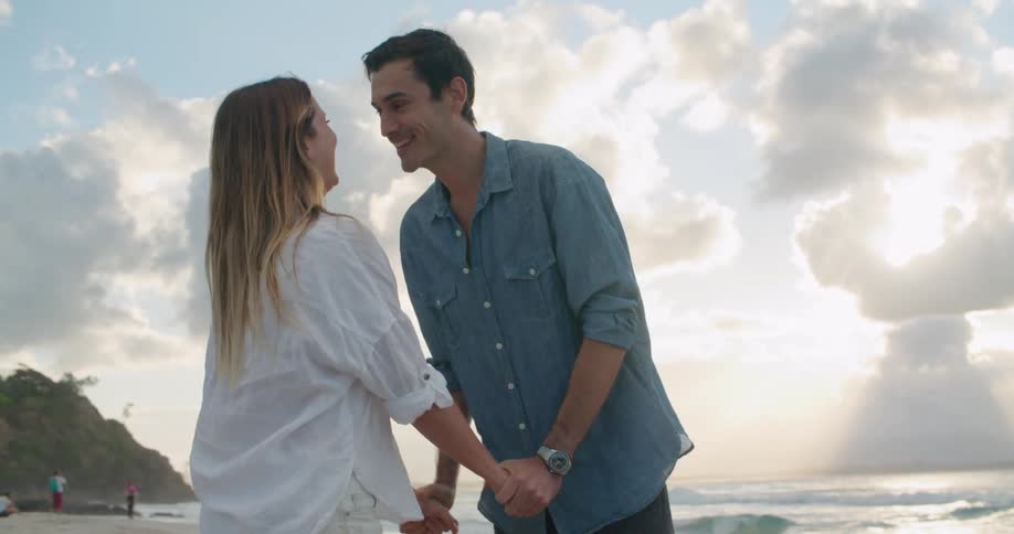 A man and a woman standing on a beach holding hands and kissing with the ocean sky and clouds in the background