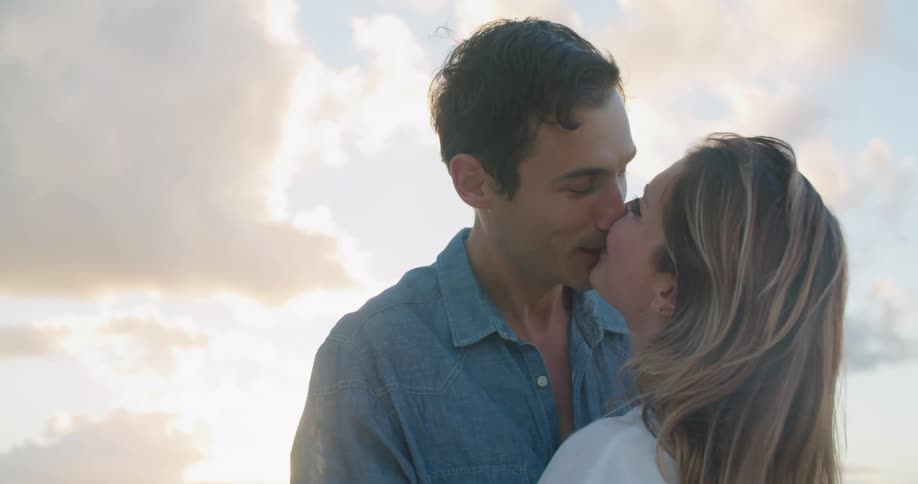 A man and woman smile as they stand close and lovingly kiss each other while outdoors