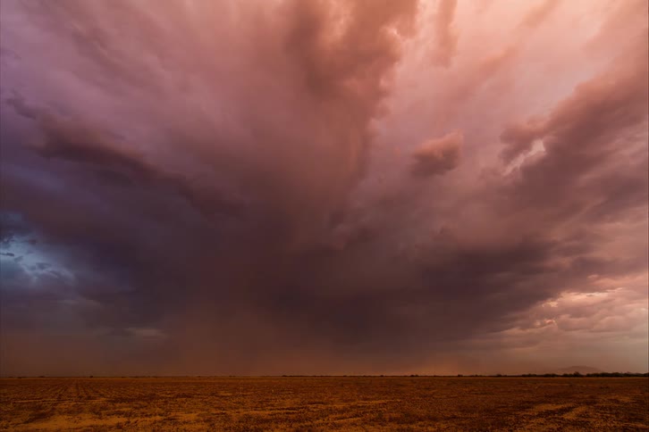 The orange sky dims as thunder clouds roll over a dusty grassland during the day