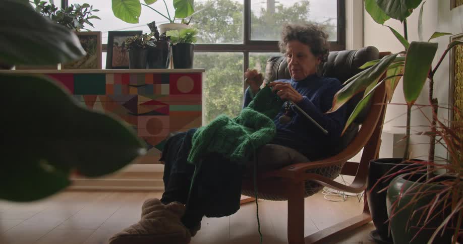 Woman wearing sweater and quilted house boots sits and knits in room decorated with houseplants