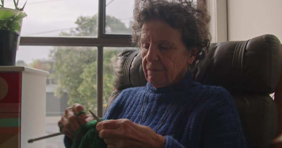 A woman knits a green blanket while sitting in a chair next to her window