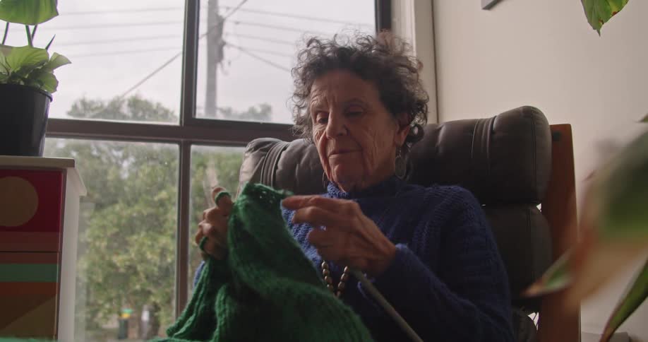 Elderly woman wearing sweater sitting in chair knits green blanket