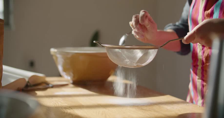 A person in a room putting flour in a strainer with a wall in the background