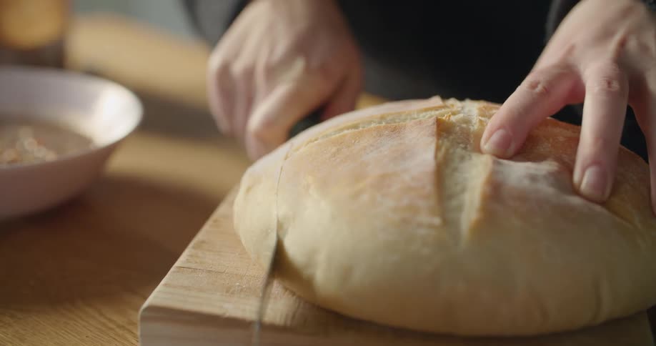 Hands hold a freshly baked round loaf of bread and slice to eat with a bowl of soup