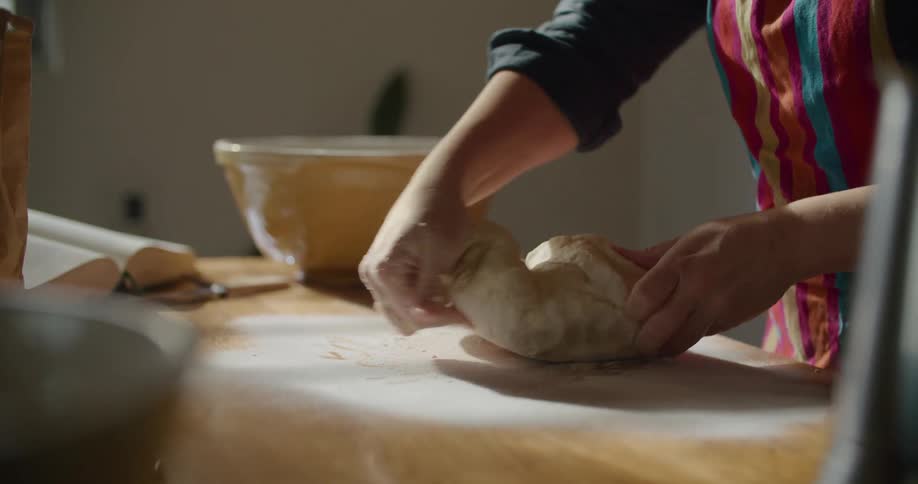 A woman places a ball of dough onto a floured wooden counter and kneads and works it