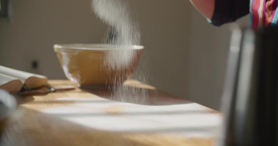 A person in a room with a strainer putting flour on a table