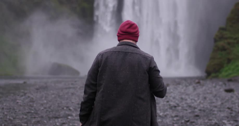 A man wearing a red beanie walks towards a waterfall