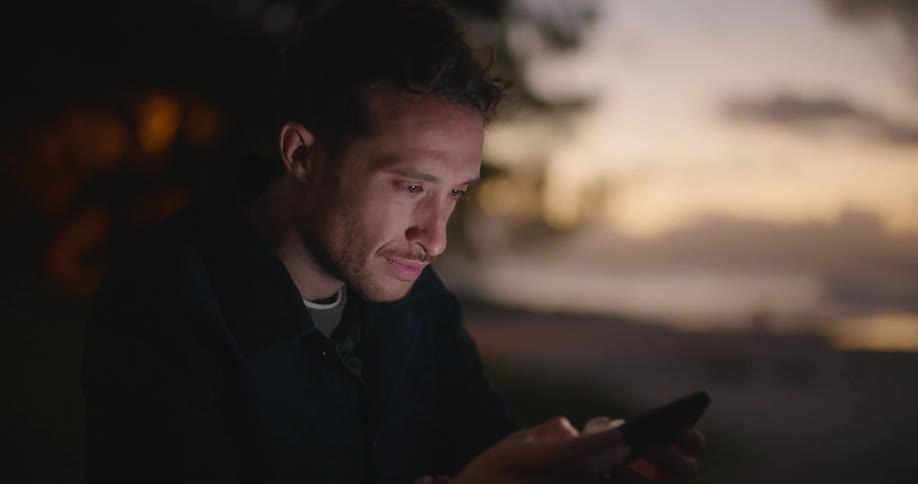 Man with a shirt and jeans sitting on hill slope uses smartphone next to the beach