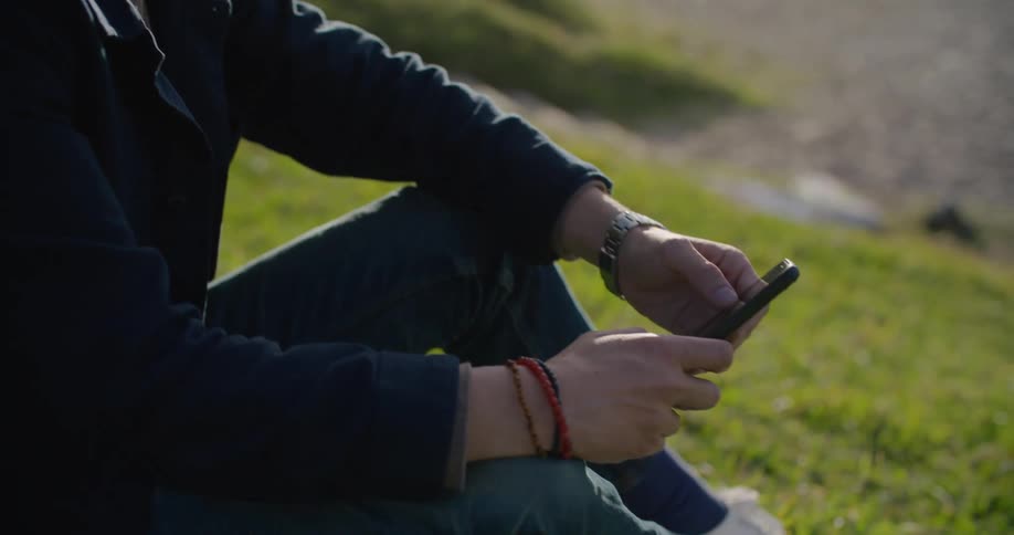 Man sits on grass near beach scrolling through mobile phone as sun reflects off water